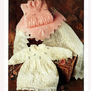 Peut inclure: Ensemble de vêtements pour bébés tricotés à la main, aux couleurs pastel douces. L'image présente une robe blanche et une rose, ainsi qu'une couverture assortie. Les robes ont des motifs complexes et sont ornées de petits éléments décoratifs. Les articles sont disposés dans un panier en osier.