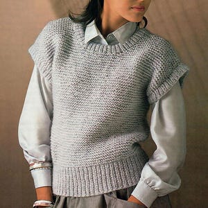 May include: A light gray knitted vest with a ribbed hem and short sleeves. The vest is worn over a light blue button-down shirt. The overall style is casual and comfortable.