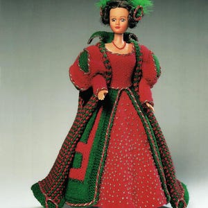 May include: A doll wearing a red and green crocheted gown with a matching cape. The gown features a red bodice and skirt with green trim and accents. The doll has dark hair with green feather accents and a red necklace.