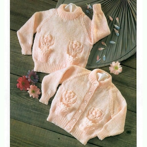 May include: Two pink knitted baby sweaters with floral designs. One sweater is a pullover and the other is a cardigan with buttons.