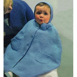 Op de afbeelding: Een baby draagt een lichtblauwe gebreide cape met bijpassende capuchon. De cape heeft twee zakken en sluit met drie knopen. Een blauwe pompon siert de capuchon. De baby kijkt recht in de camera.