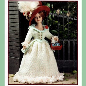 Puede incluir: Una muñeca con un vestido blanco de ganchillo con un dobladillo con volantes y un sombrero rojo de ganchillo con un adorno de plumas blancas. La muñeca lleva una cesta roja con una tapa de ganchillo blanca.
