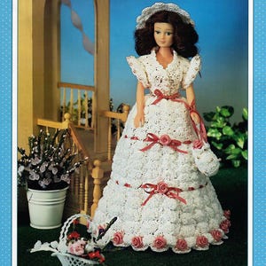 May include: A doll wearing a white crocheted dress with a matching hat and purse. The dress features red ribbon accents and rose embellishments. The doll is standing in front of a porch with a white picket fence and a potted plant.