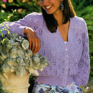 May include: A light purple knitted cardigan with a V-neck and button closure. The cardigan has a delicate lace pattern and is worn over a floral print skirt.