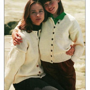 Puede incluir: Dos personas con cárdigans de punto color crema a juego con botones oscuros. Los cárdigans tienen un dobladillo y puños acanalados. Una persona lleva pantalones oscuros y la otra pantalones de pana marrones. El fondo es una escena borrosa al aire libre.