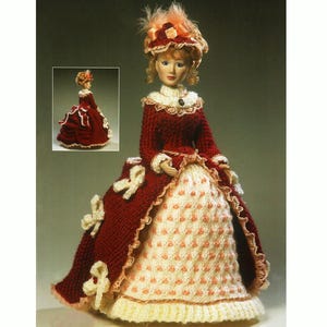 Puede incluir: Una muñeca de punto que lleva un vestido rojo y blanco con ribete de encaje. La muñeca lleva un sombrero blanco con una rosa roja y plumas.