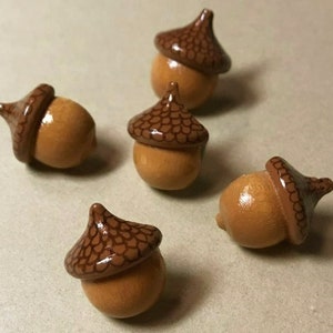 May include: Five wooden acorns painted with a brown and tan pattern. The acorns are approximately 1 inch in size.
