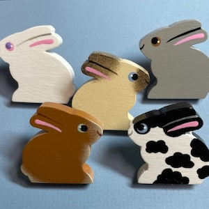 May include: Five wooden rabbit figurines painted in different colors. The rabbits are white, gray, brown, beige, and black and white. They are all sitting with their ears up.