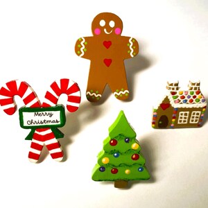 May include: A set of four Christmas-themed wooden ornaments including a brown gingerbread person, a green Christmas tree with colorful ornaments, a gingerbread house with a red heart, and two red and white candy canes with a sign reading Merry Christmas.