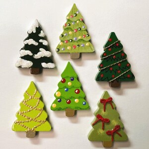 May include: Six flat, decorative wooden Christmas tree ornaments in various shades of green. Designs include a snow-covered tree, a tree with colorful painted lights, a tree with red ornaments and gold garland, a tree with gold string, and a tree with red bows.