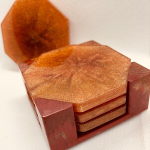 May include: A set of four octagonal resin coasters in shades of copper and gold, with a matching holder. The coasters have a shimmering, marbled effect, and the holder is a deep reddish-brown.