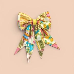 May include: A large, colorful bow made of patterned fabric. The bow features a mix of floral, striped, and geometric designs in shades of yellow, pink, blue, and green. The bow is tied with a yellow band.