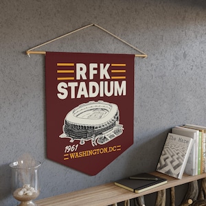 May include: A maroon wall banner featuring a white line drawing of RFK Stadium. The text reads RFK STADIUM in large white letters with yellow horizontal lines above and below. Below the stadium graphic is the text 1961 WASHINGTON, DC in yellow.