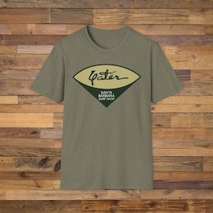 May include: An olive green short-sleeve t-shirt featuring a graphic on the chest. The graphic is a dark green and tan diamond shape containing the cursive text "Yater" above the words "SANTA BARBARA SURF SHOP" printed in white capital letters.