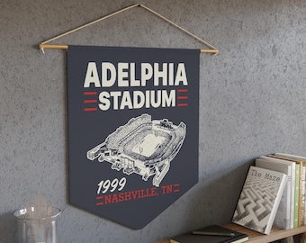 以下が含まれることがあります： ダークグレーの壁掛けバナー。白い文字で「ADELPHIA STADIUM」、白い線画のスタジアム、赤い文字で「1999 NASHVILLE, TN」が描かれています。バナーは木製の棒と明るい色の紐で、質感のあるグレーの壁に掛けられています。