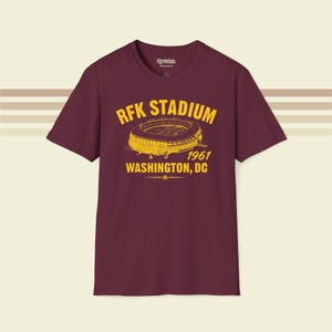 May include: A maroon short-sleeve t-shirt featuring a yellow graphic of RFK Stadium. The text reads RFK STADIUM, 1961, and WASHINGTON, DC. The design includes a stylised illustration of the stadium architecture centred on the chest area.