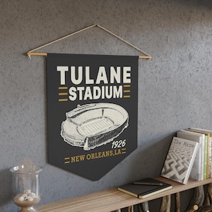 May include: A dark gray wall banner featuring a white illustration of Tulane Stadium. The text reads TULANE STADIUM, 1926, and NEW ORLEANS, LA, with gold horizontal lines. The banner hangs from a wooden dowel with a light-colored string.