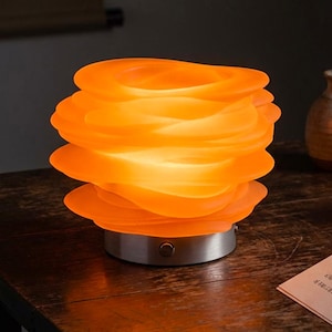 May include: A glowing orange table lamp with a layered, wavy, translucent shade resting on a circular silver metal base. The lamp emits a warm light, highlighting the undulating, organic curves of the stacked glass-like material.
