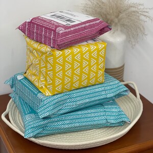 May include: A stack of three parcels wrapped in patterned paper. The top parcel is pink with white polka dots and the text "Midwest Makers" printed on it. The middle parcel is yellow with a white chevron pattern. The bottom parcel is teal with a white chevron pattern. The parcels are sitting on a white rope basket.