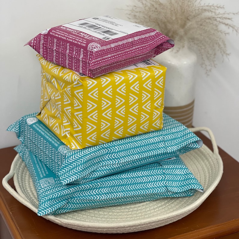 May include: A stack of three parcels wrapped in patterned paper. The top parcel is pink with white polka dots and the text "Midwest Makers" printed on it. The middle parcel is yellow with a white chevron pattern. The bottom parcel is teal with a white chevron pattern. The parcels are sitting on a white rope basket.