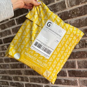 May include: A yellow padded mailing envelope with a white geometric pattern. The envelope has a white label with the text "USPS GROUND ADVANTAGE" and a barcode. The envelope also has a label with the text "MIDWEST MAILERS - Sustainably Stylish" and a QR code.