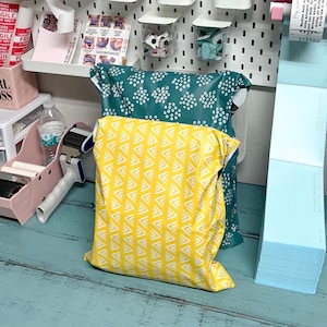 May include: Two poly mailers, one yellow with a white geometric pattern and one teal with white polka dots. The mailers are stacked on top of each other on a teal wooden surface.