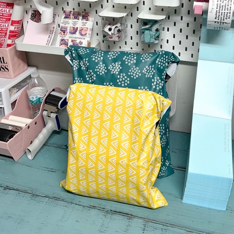May include: Two poly mailers, one yellow with a white geometric pattern and one teal with white polka dots. The mailers are stacked on top of each other on a teal wooden surface.