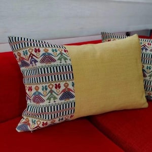 Può includere: Cuscino decorativo rettangolare con un centro giallo e un'estremità a fantasia. L'estremità a fantasia presenta un disegno di figure con abiti colorati, su uno sfondo a righe bianche e nere. Il cuscino è su una superficie rossa.