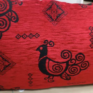 Könnte beinhalten: Rotes Kissen mit einem schwarzen, stilisierten Vogeldesign. Der Vogel hat einen Kamm und einen langen Schwanz mit Wirbeln. Das Kissen hat ein geometrisches Muster in Schwarz.