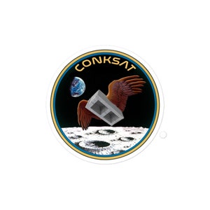 May include: A round sticker with a space-themed design. The sticker features the text "CONKSAT" in gold lettering, a concrete block with brown wings, a blue and white planet, and a lunar surface.
