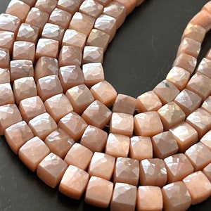 May include: A strand of iridescent pink sunstone beads, each bead is faceted and shaped like a square. The beads are strung together on a clear thread.