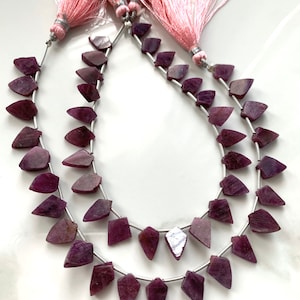 May include: A strand of purple gemstone beads with a faceted, triangular shape. The beads are strung on a silver wire and have a pink tassel at each end.