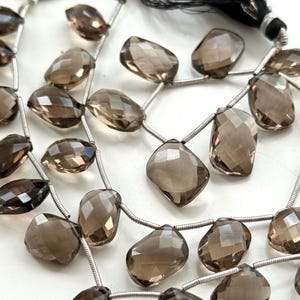 May include: Strands of faceted, smoky quartz gemstones. The beads are a translucent brown color and are strung on silver-colored wire. The beads have a variety of shapes, including teardrops and rectangles. A black tassel is visible in the background.