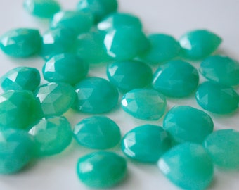 May include: A collection of teal green gemstone cabochons, each with a faceted, oval shape. The stones are arranged in a loose pile on a white surface.