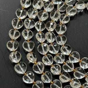 May include: A close-up of a necklace featuring clear, heart-shaped beads strung together with gold-colored connectors. The beads are faceted, catching the light and creating a sparkling effect. The necklace is displayed against a dark background.