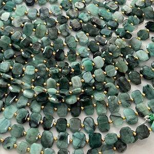 May include: A strand of emerald green gemstone beads with a faceted, rectangular shape. The beads are strung on gold-colored wire.