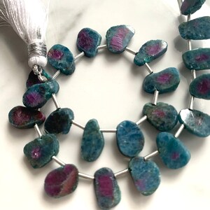 May include: A strand of blue and pink gemstone beads with a white tassel. The beads are irregularly shaped and have a natural, rough texture.