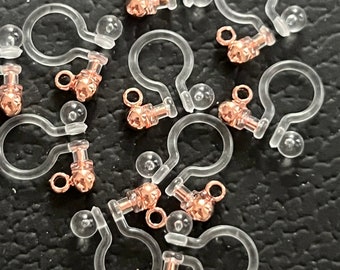 May include: A group of clear plastic earring backs with rose gold coloured metal posts and balls. The backs are designed to be used with pierced earrings.