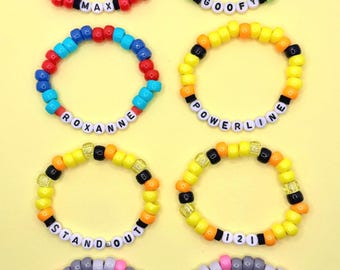 Op de afbeelding: Zes kleurrijke kralenarmbanden met verschillende namen erop gespeld. De namen zijn Max, Goofy, Roxanne, Powerline, Stand Out en 121. De armbanden liggen op een gele achtergrond.