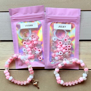 May include: Two pink zippered bags with clear windows, each containing a set of pink, white, and clear beads, a gold ballerina charm, and a white elastic string. The bags are labeled "Vivian" and "Juliet".