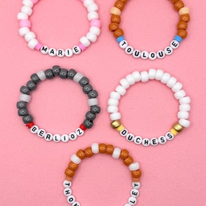 May include: Five beaded bracelets with names spelled out in white letter beads. The bracelets feature various color combinations, including pink and white, brown and tan, gray and white, white and gold, and orange and white. The names on the bracelets are "Marie", "Toulouse", "Berlioz", "Duchess", and "Thomas O'Malley".
