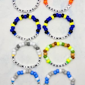 May include: A collection of colorful beaded bracelets with various phrases. The bracelets feature a variety of colors, including red, orange, blue, yellow, green, and white. Each bracelet has a different phrase spelled out in white letter beads, such as "BEYOND THE SEA" and "SHARK BAIT".