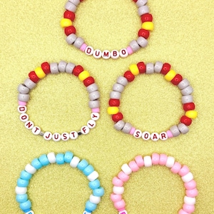 May include: Five beaded bracelets in various colors. One bracelet says "DUMBO" in white letter beads. Other bracelets say "DON'T JUST FLY", "SOAR", "BABY MINE", and "BABY MINE" in white letter beads. The bracelets are on a gold background.