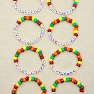 May include: Eight colorful beaded bracelets with various phrases. The bracelets feature a mix of yellow, green, orange, brown, and red beads. Each bracelet has white letter beads spelling out phrases like "TIKI TIKI TIKI", "TIKI ROOM", and "ALL THE BIRDS".