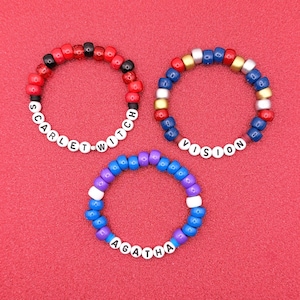 May include: Three beaded bracelets with white letter beads spelling "SCARLET WITCH", "VISION", and "AGATHA". The bracelets feature red, black, blue, gold, and purple beads. The background is a solid red color.