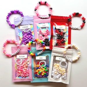 May include: A collection of colorful beaded bracelets with names spelled out in white letter beads. The bracelets are in various colors, including pink, purple, red, and rainbow. Small bags of beads in matching colors are also visible.