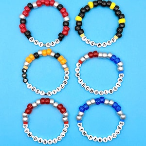 May include: Six colorful beaded bracelets with white letter beads spelling out "ANT-MAN", "THE WASP", "TEST KITCHEN", "PYM TECHNOLOGIES", and "PYM PARTICLES". The bracelets are on a blue background.