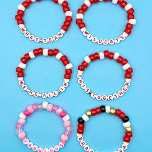 May include: Six beaded bracelets in red, white, pink, and gold, with text. The bracelets say "WHAT TEAM", "WILDCATS", "WE'RE ALL IN", "THIS TOGETHER", "FABULOUS", and "BRAVO BRAVA". The bracelets are on a blue background.