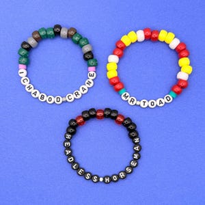 May include: Three beaded bracelets on a blue background. One bracelet is green, brown, and black with the text "ICHABOD CRANE". Another is red, yellow, and white with "MR. TOAD". The third is black and red with "HEADLESS HORSEMAN".