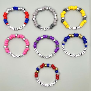 May include: Seven colorful beaded bracelets. Each bracelet features a different color scheme, including red, blue, yellow, pink, gray, and purple. Some bracelets have white letter beads spelling out names like "ORION PAX" and "STARSCREAM".